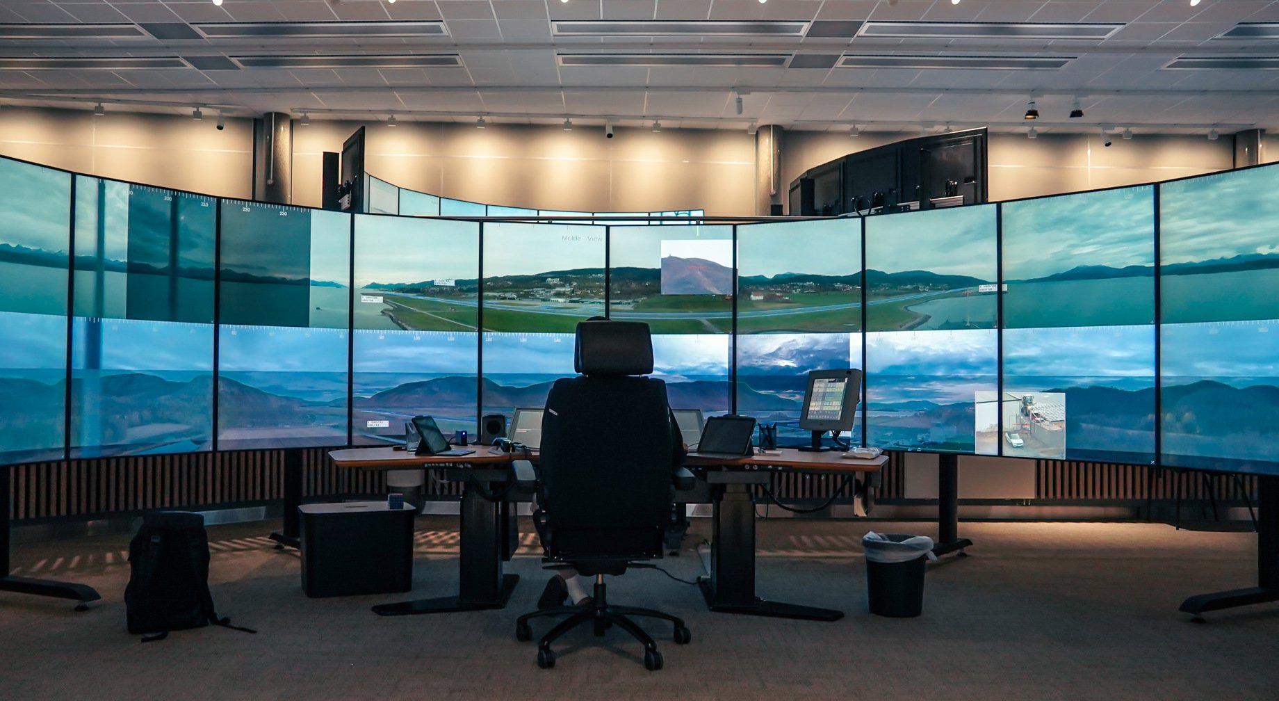 Stor luftfartskontrollrom med flere skjermer som viser flyplasser og luftutsikt. Sentral pult med stol, skjermer og utstyr under taklys i et høyteknologisk miljø