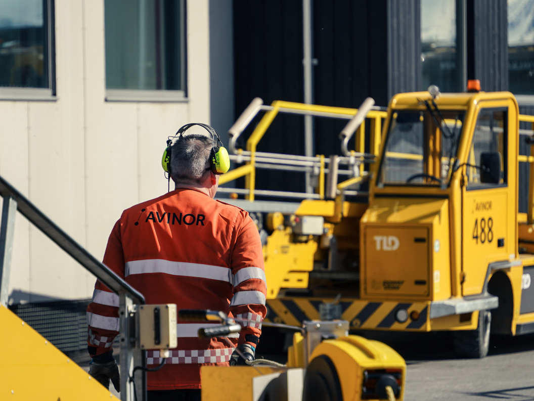 Flyplassbakkemannskapsarbeider iført oransje sikkerhetsutstyr og støydempende hodetelefoner, som står nær et gult flyplasstjenestekjøretøy på en solrik dag
