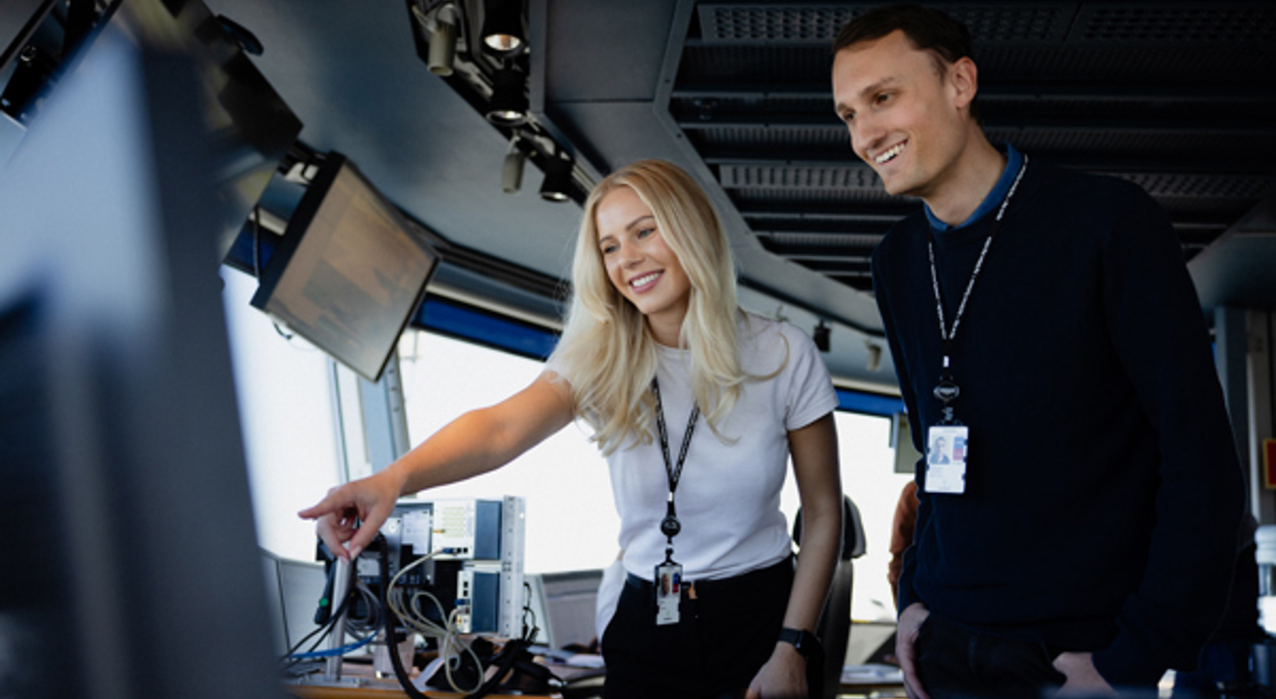 To fagfolk i et kontrollrom som diskuterer over en dataskjerm, omgitt av teknisk utstyr og skjermer, som viser samarbeid og teknologi