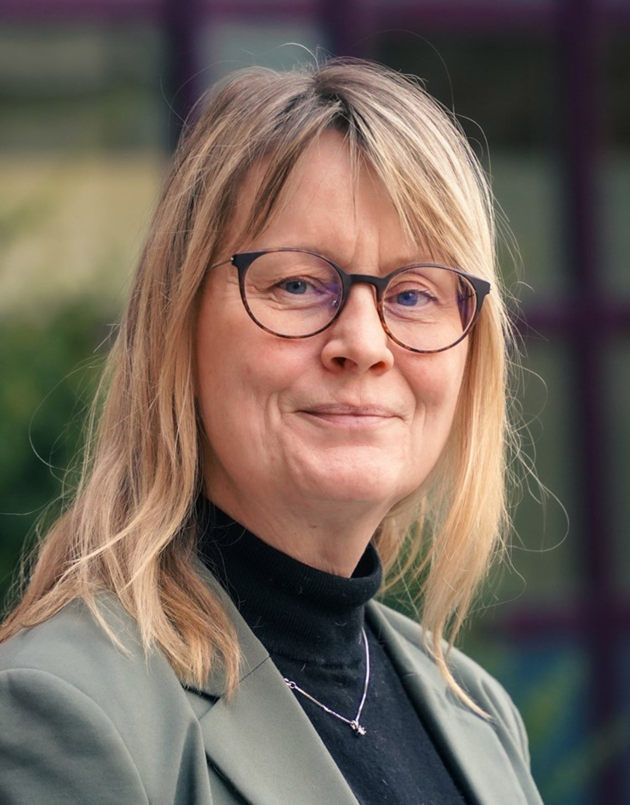 En kvinne med briller og blondt hår smiler i et portrettbilde. Hun har på seg en olivengrønn jakke og sort høyhalset genser, med en uskarp bakgrunn