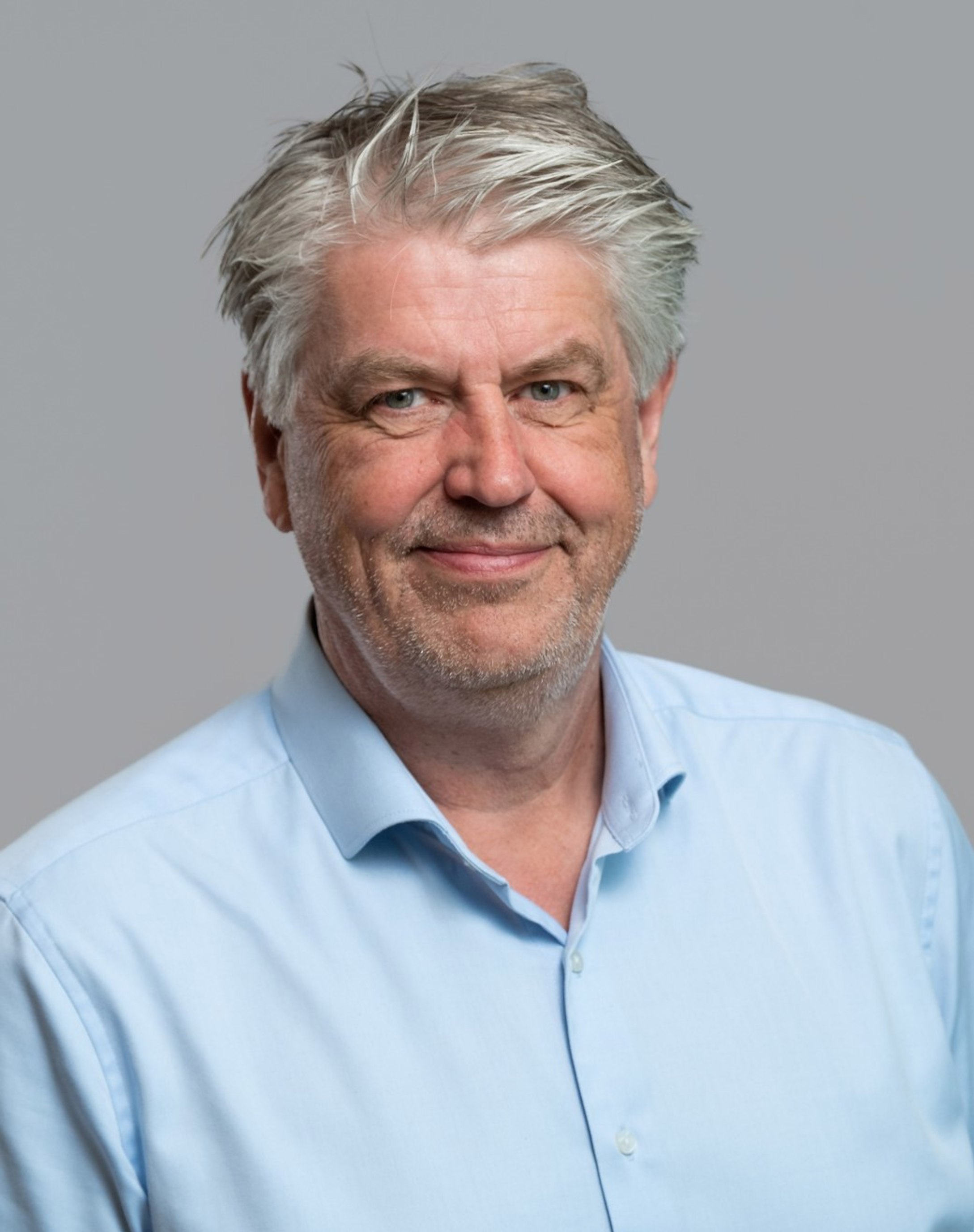 Eldre mann med grått hår og skjegg iført en lys blå skjorte, ser vennlig ut mot kameraet. Grå bakgrunn