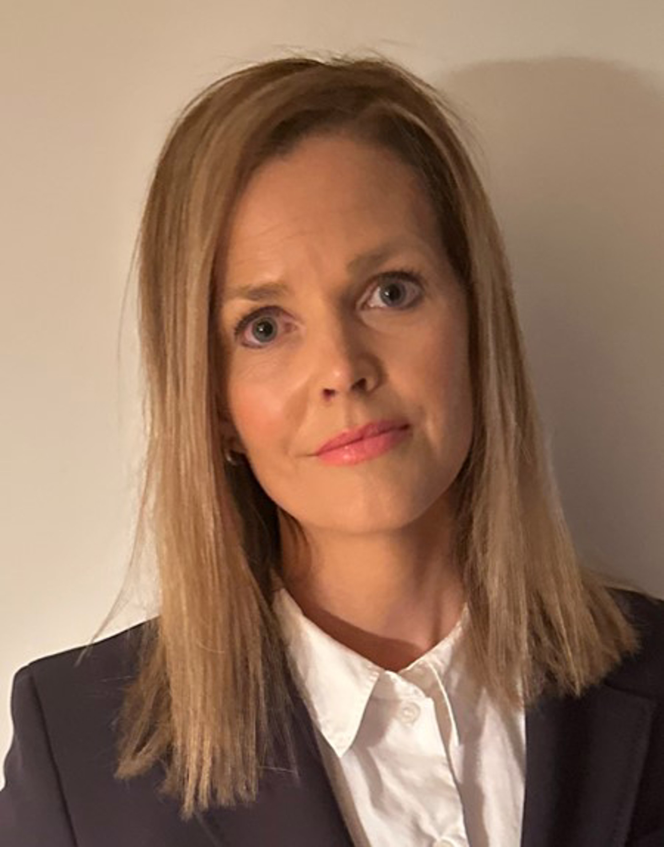 Kvinne med rett, skulderlangt blondt hår iført en mørk blazer og hvit skjorte mot en nøytral bakgrunn