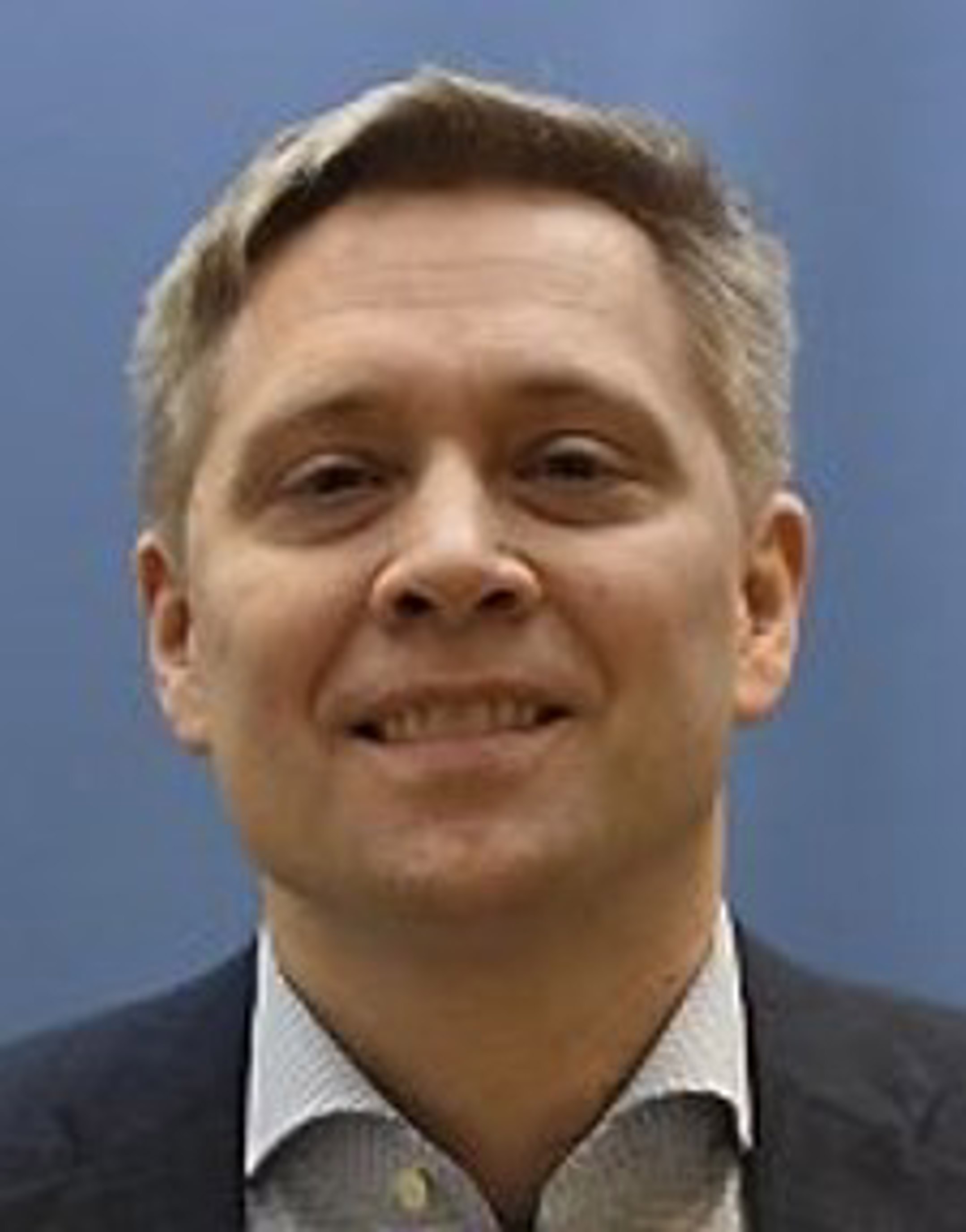 Mann i dress med blå bakgrunn, smiler til kameraet