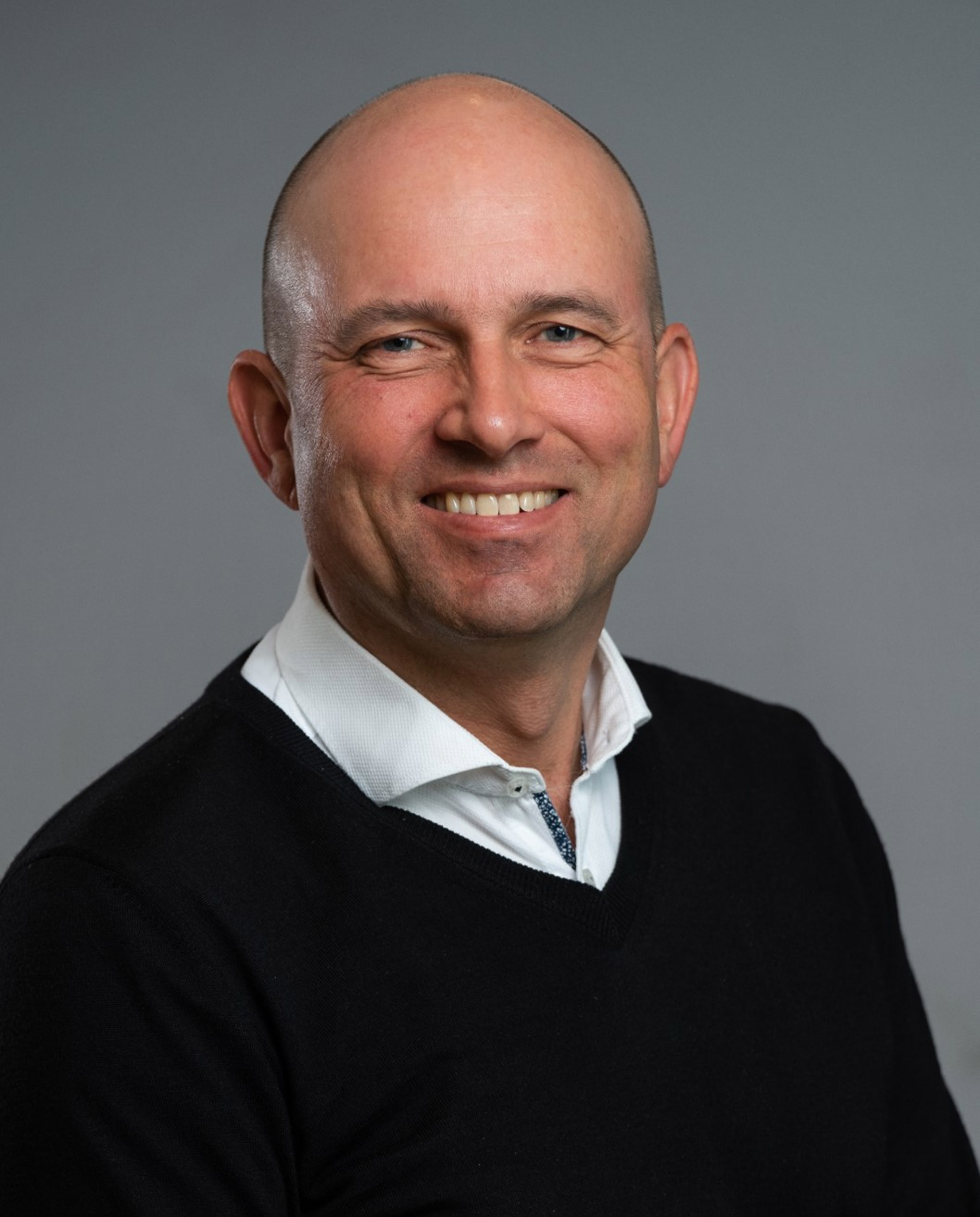 Smilende skallet mann i en hvit skjorte med krage og svart genser mot en grå bakgrunn