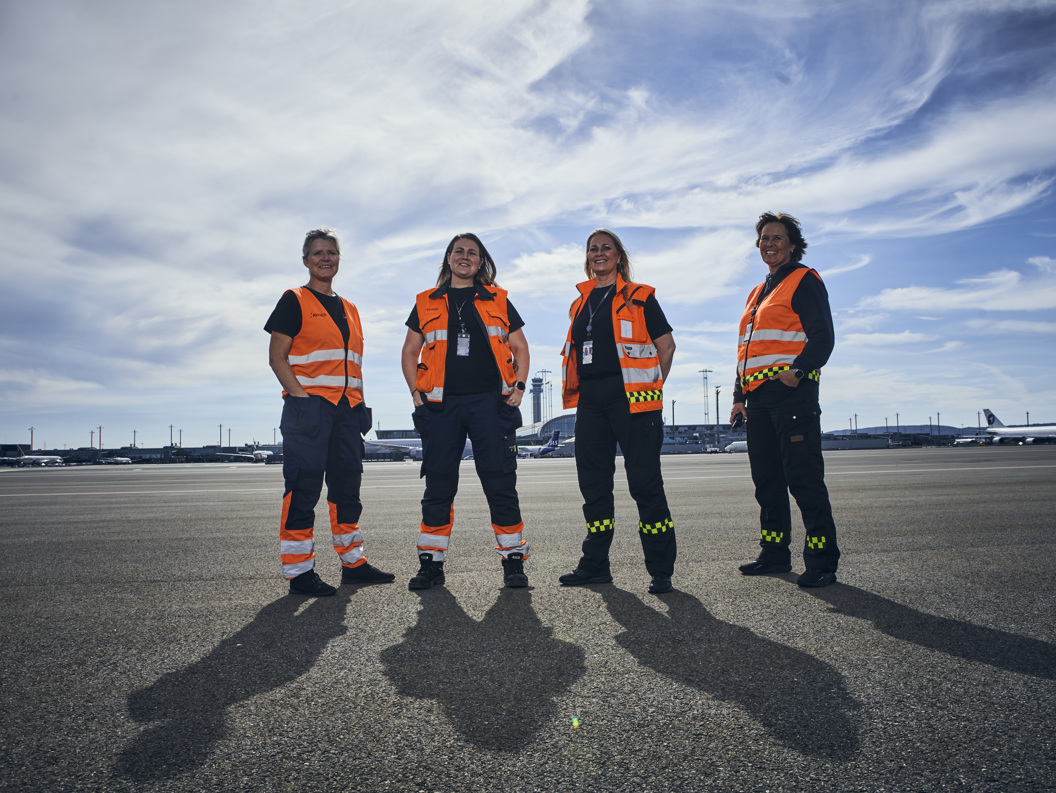 Four airport ground crew members wearing orange safety vests standing on a tarmac with airplanes and control towers in the background under a cloudy sky