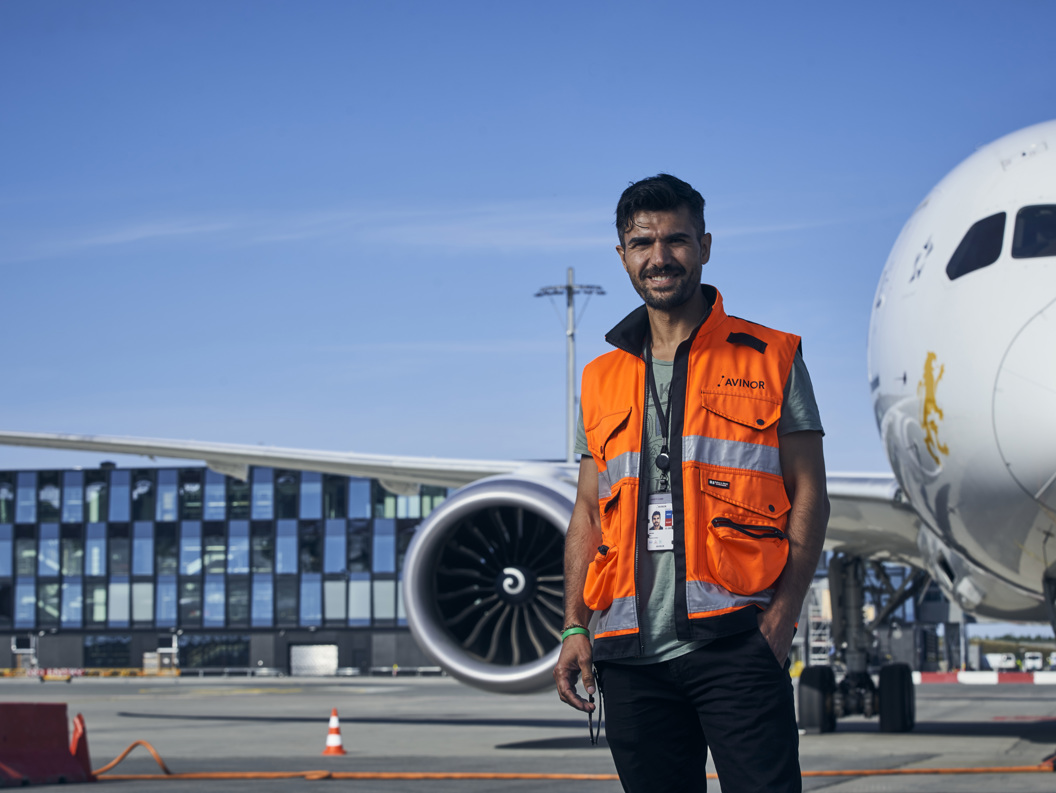 Flyplassarbeider i oransje sikkerhetsvest står selvsikkert ved siden av et parkert fly på en solfylt dag, med terminalbygningen i bakgrunnen