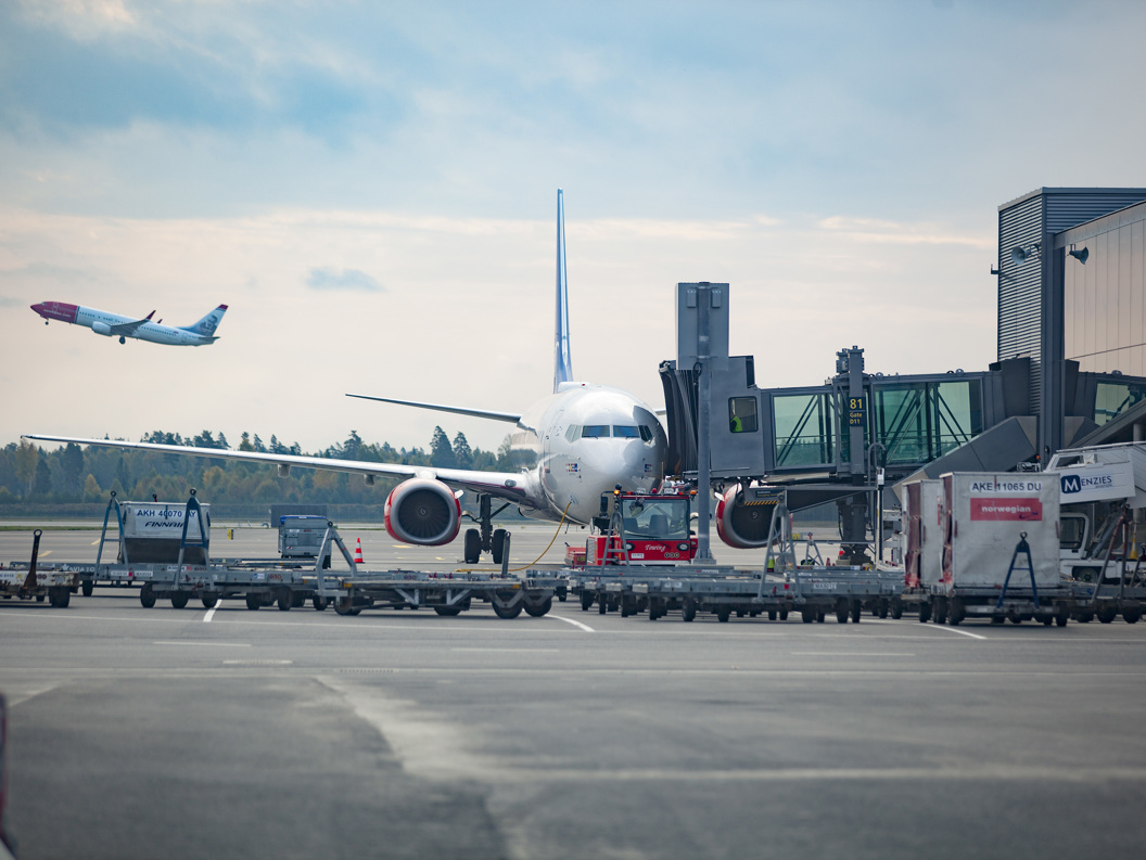 Fly parkert ved en flybro på en flyplass med et annet fly som tar av i bakgrunnen mot en skyet himmel