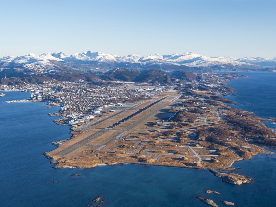 Luftfoto av Bodø i Norge med Bodø lufthavn i forgrunnen og snødekte fjell i bakgrunnen. Byen ligger langs kystlinjen med det blå havet på begge sider av halvøya.