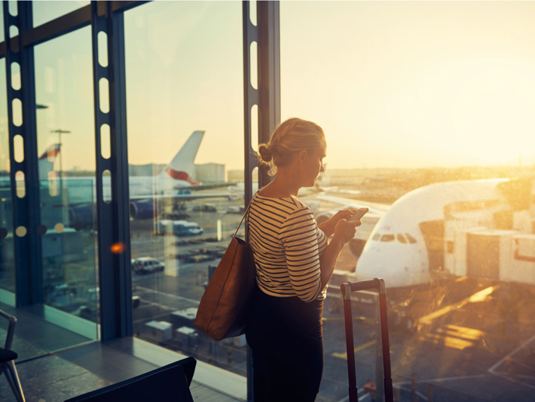 Reisande brukar smarttelefon på flyplassterminal med fly i bakgrunnen under solnedgang
