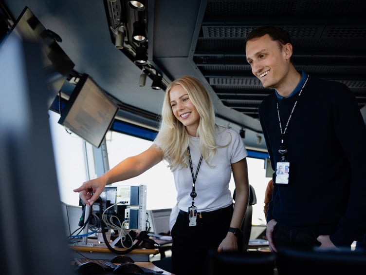 To personer i et kontrollrom som ser på en skjerm, smilende og engasjerte i arbeidet sitt.