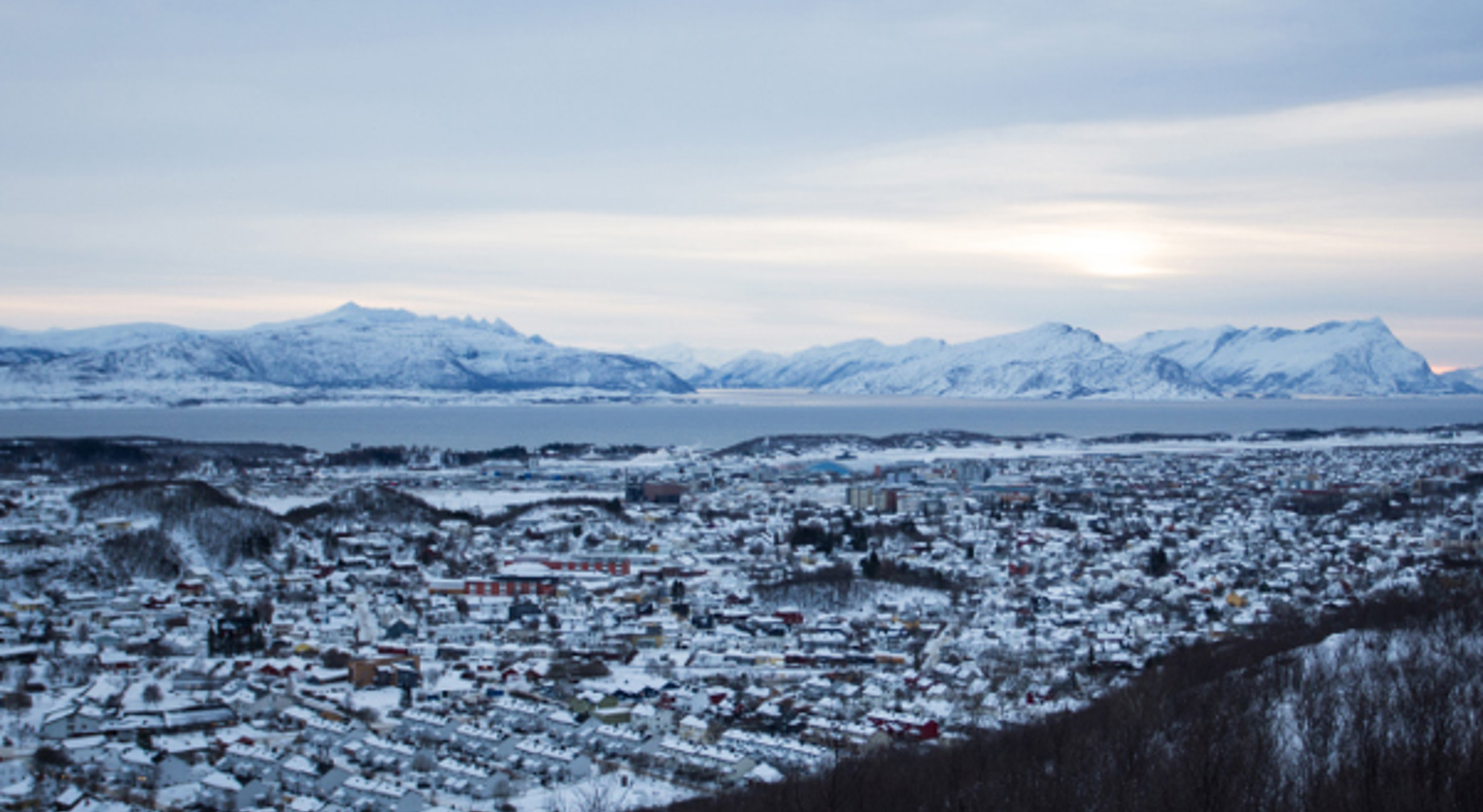 Vinterlandskap i Noreg med snødekte fjell, hus, og ein fjord i bakgrunnen under ein overskya himmel.