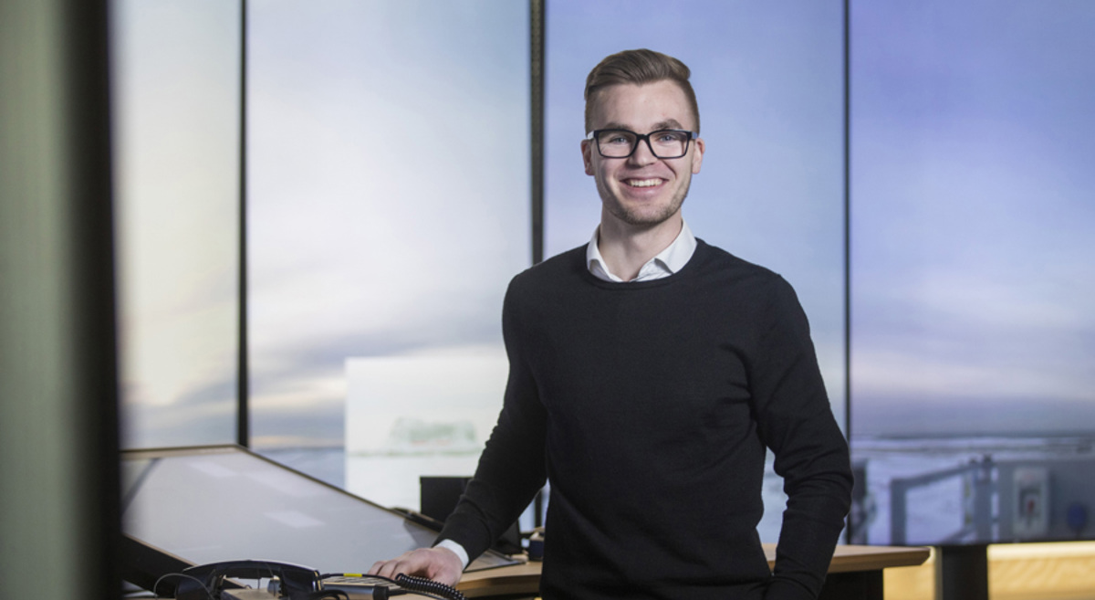 Ein smilande person med briller står ved eit skrivebord med teknologiutstyr, mot ei moderne kontorinnstilling.