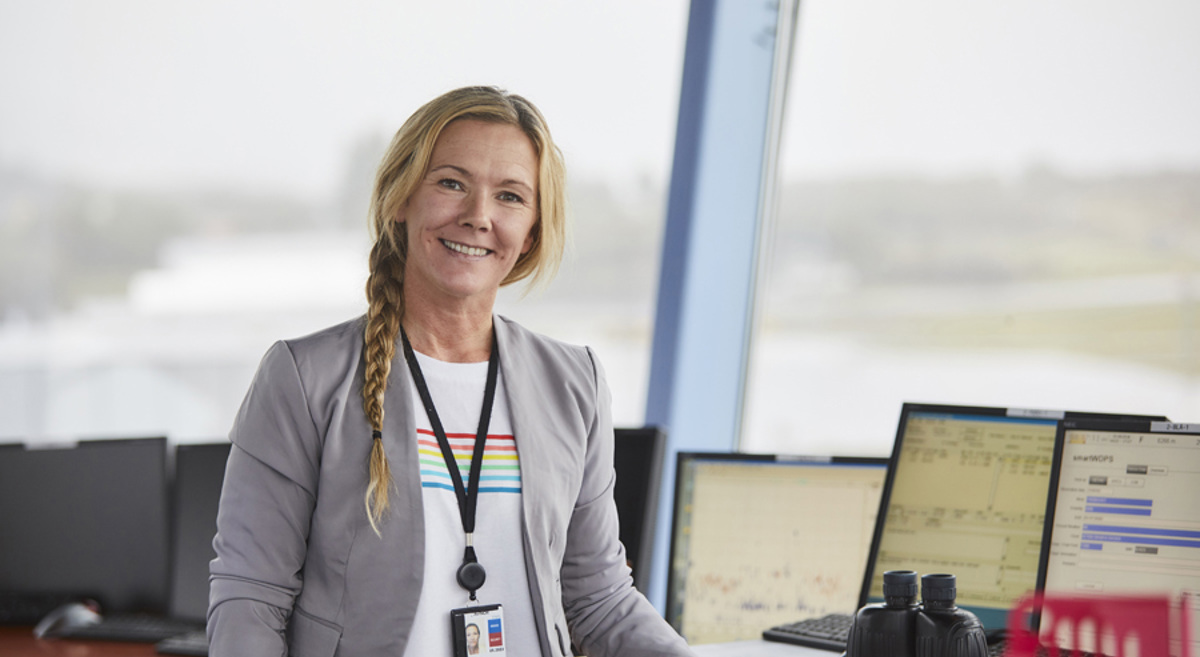 Kvinne i kontrollrom med dataskjermar i bakgrunnen, iført grå jakke og ID-kort.