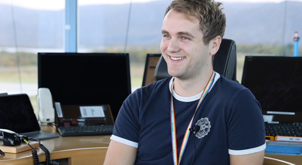 En ung mann sitter smilende ved et moderne arbeidsområde med flere dataskjermer i bakgrunnen, iført en mørkeblå t-skjorte og regnbuefarget lanyard. Bakgrunnen viser et landskap gjennom vinduet.