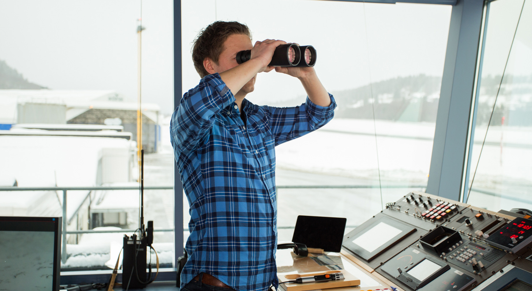Mann i blå rutete skjorte bruker kikkert i eit kontrollrom med fleire skjermar og kontrollpanel. Utsikt over snødekt landskap gjennom vindauga.