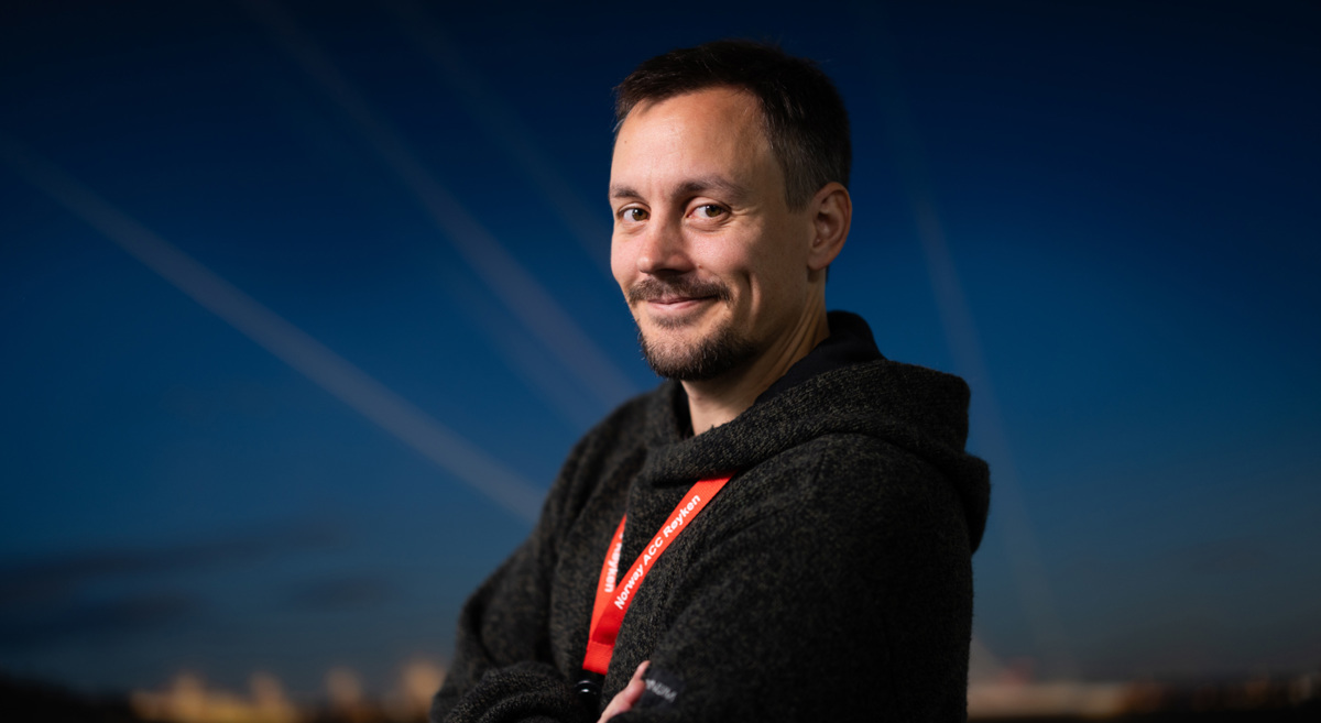 Mann i mørk genser står utendørs med armene i kors, smiler og ser mot kameraet. Bakgrunnen er en klar nattehimmel med lysstriper.