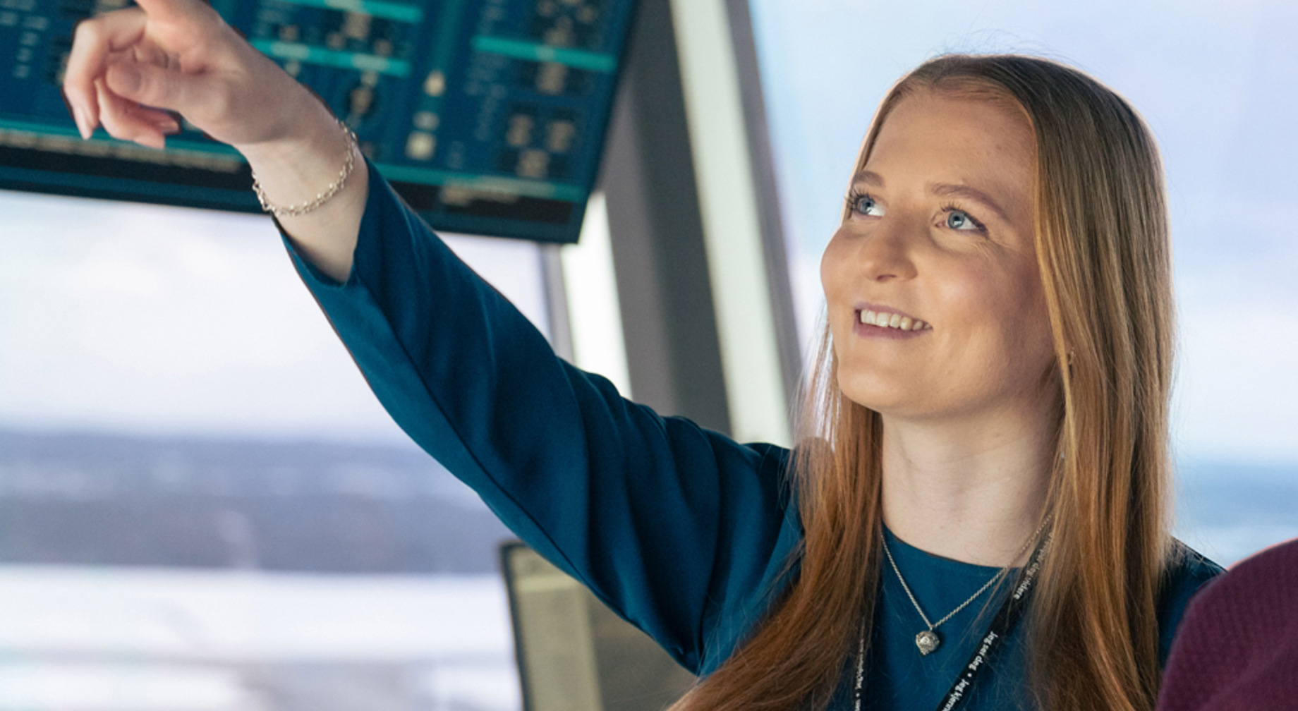 Kvinne peker mot skjerm i kontrollrom, smiler og ser fokusert ut. Skjermer med data i bakgrunnen.