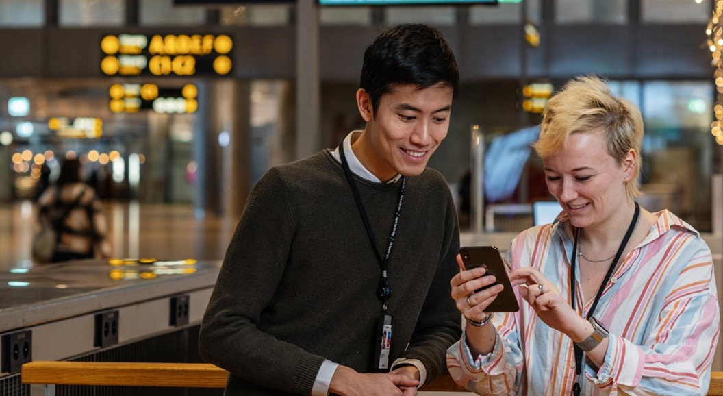 To personer ser på en mobiltelefon på en moderne flyplass, med skilt og informasjonstavler i bakgrunnen.