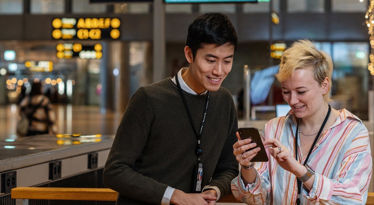 To personer ser på en mobiltelefon på en moderne flyplass, med skilt og informasjonstavler i bakgrunnen.
