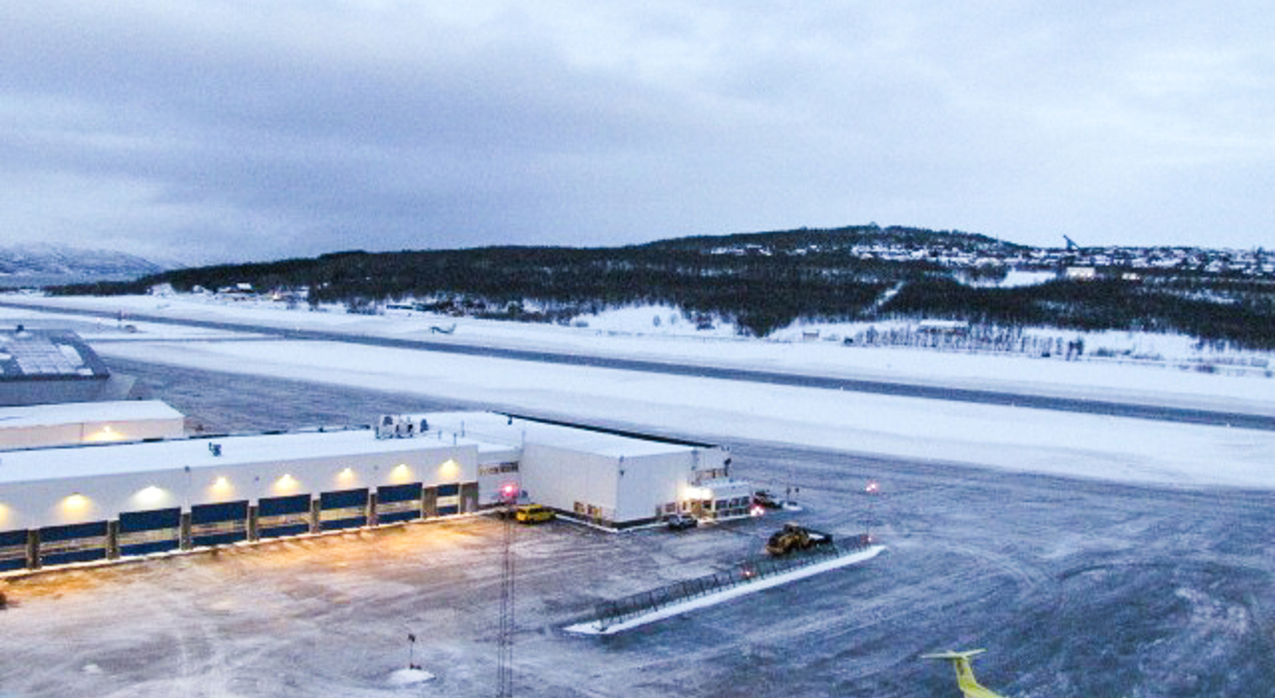 Flyplass med snødekt rullebane ved daggry, gult fly parkert ved siden av hangar.