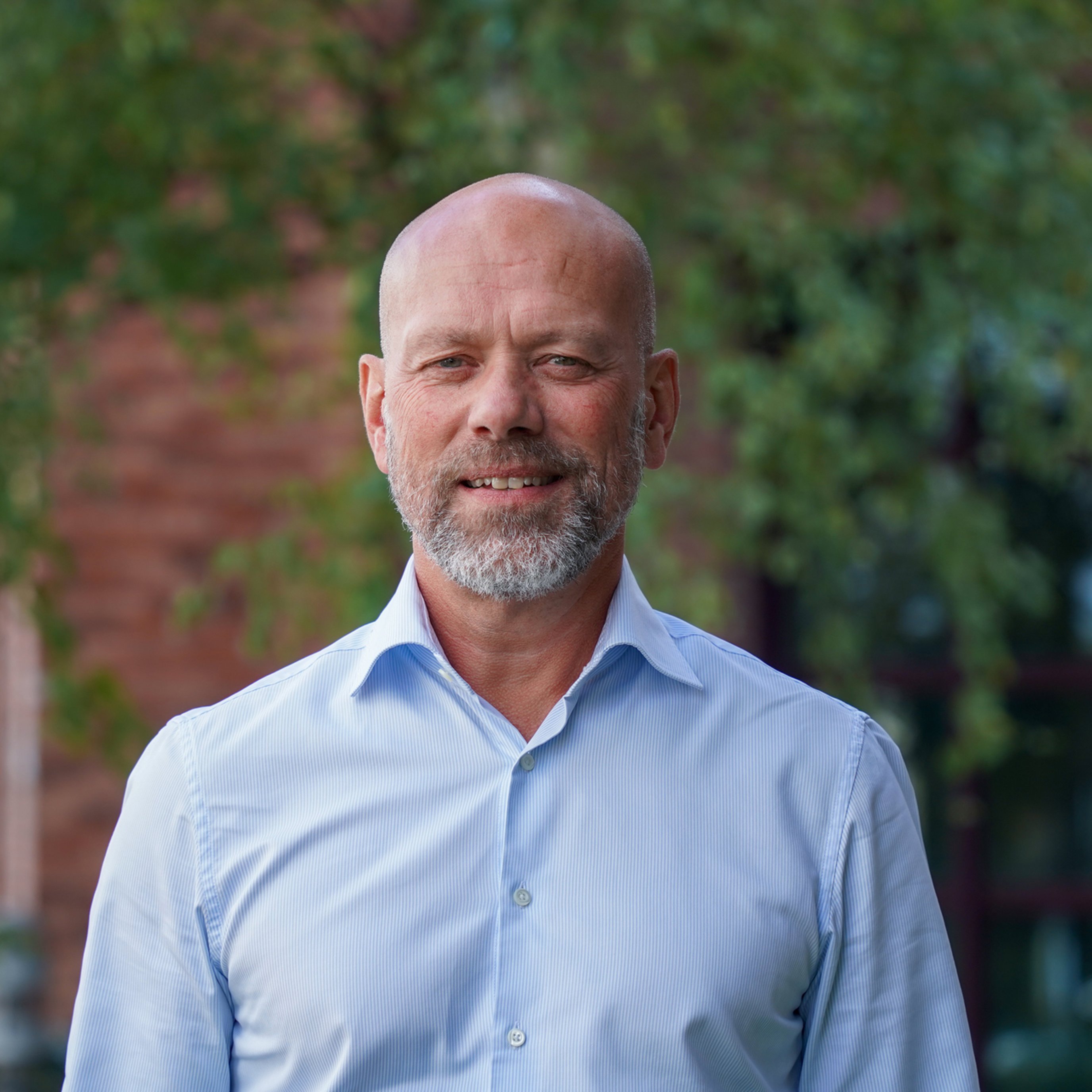 En smilende mann med skjegg og blå skjorte står ute foran en grønn bakgrunn.