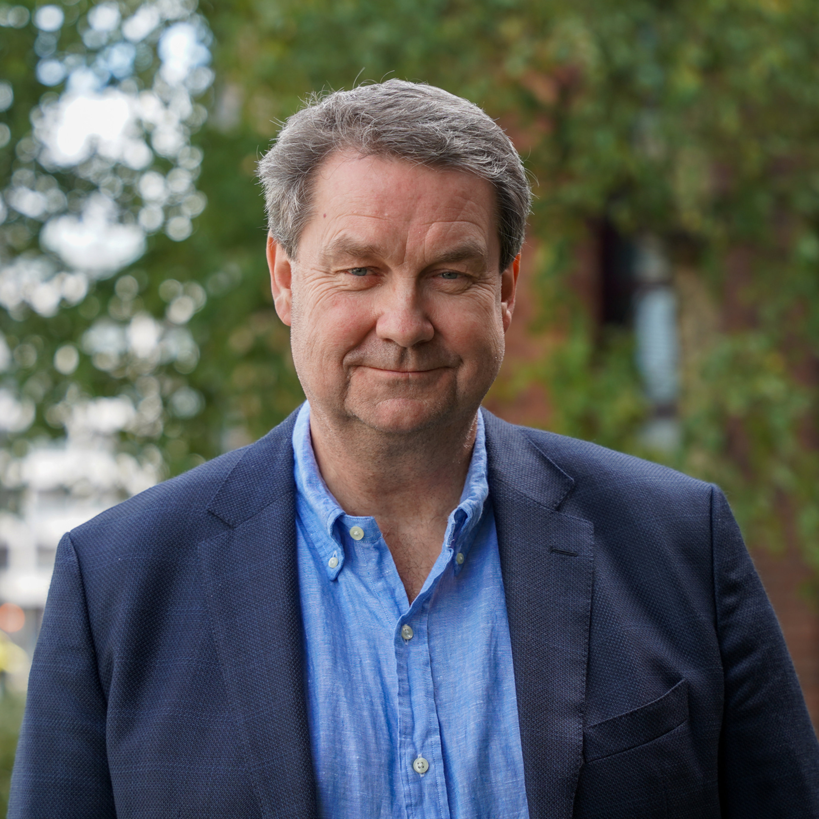 En mann i blå skjorte og blazer står utendørs foran grønne trær.