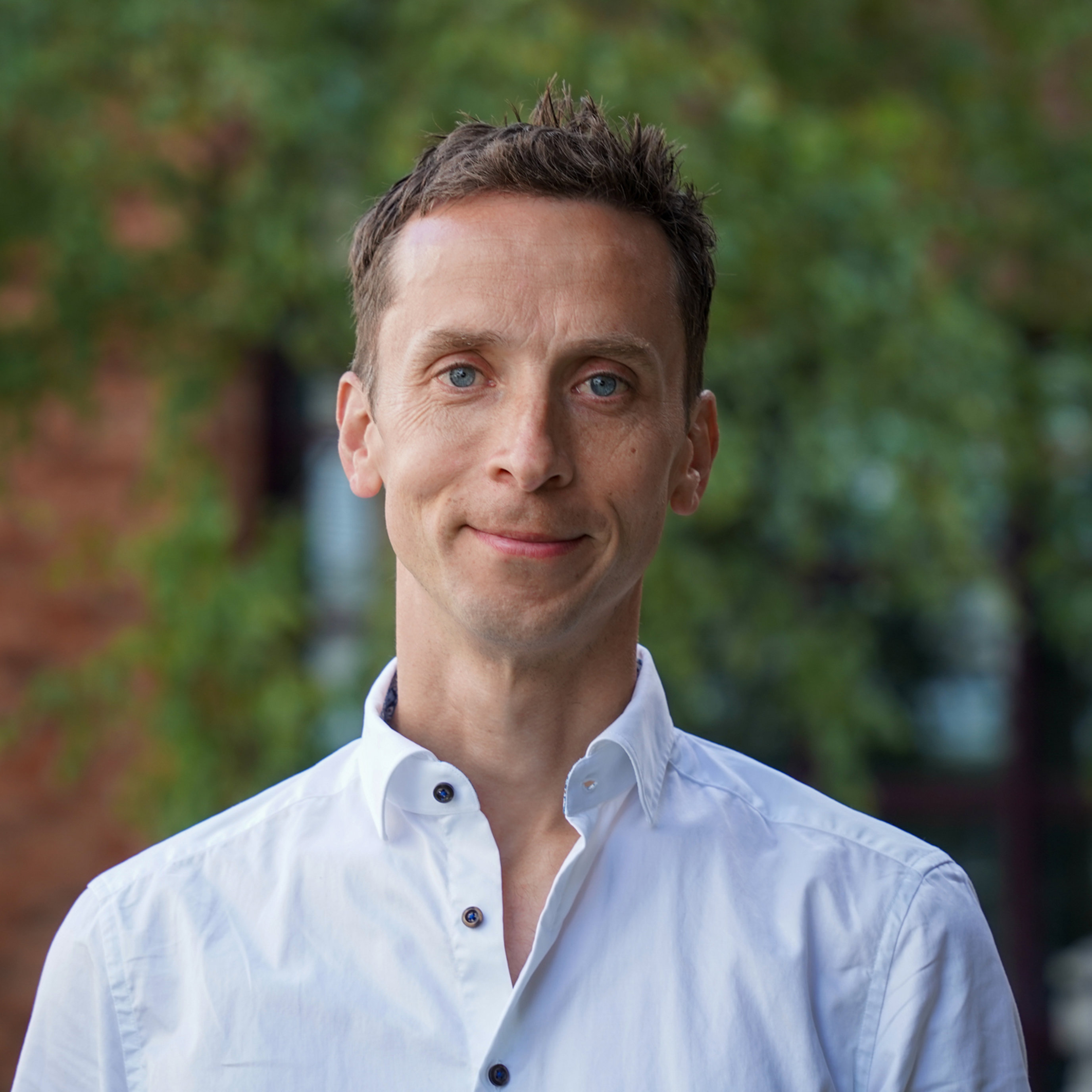 Mann i hvit skjorte med bakgrunn av grønt løv og murbygning.