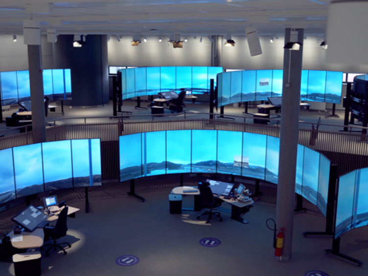 Kontrollrom med store, buede skjermer som viser landskapsbilder, flere arbeidsstasjoner med datamaskiner og kontorstoler, LED-belysning i taket.
