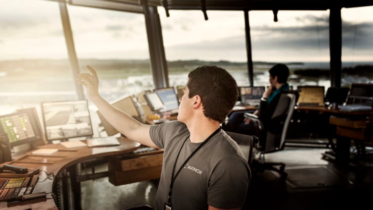 Mann i tårnkontroll med skjermer, peker ut av vinduet mens en kollega jobber i bakgrunnen på en flyplass, solnedgang i horisonten.