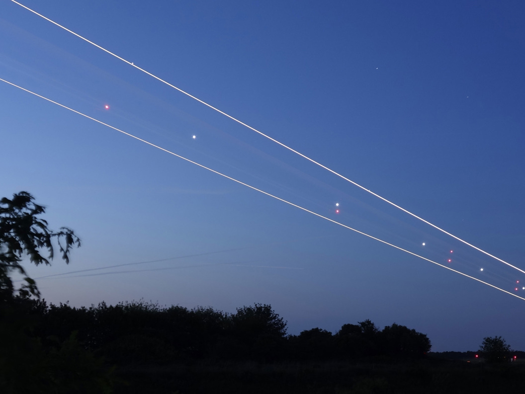 Langtidseksponeringsfoto av nattehimmelen med flylysstriper og uskarpe trær i forgrunnen, mot en stadig mørkere skumringsbakgrunn