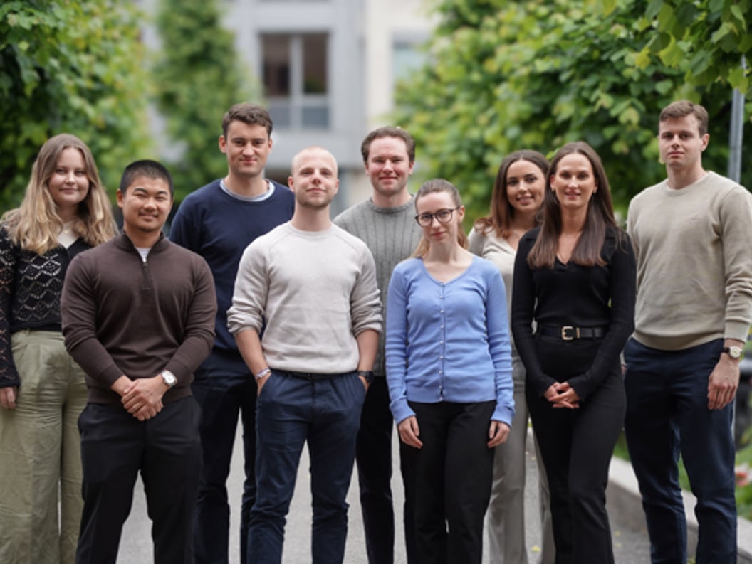 A diverse group of nine young professionals standing in an outdoor setting, posing confidently and smiling, with trees and a building in the background